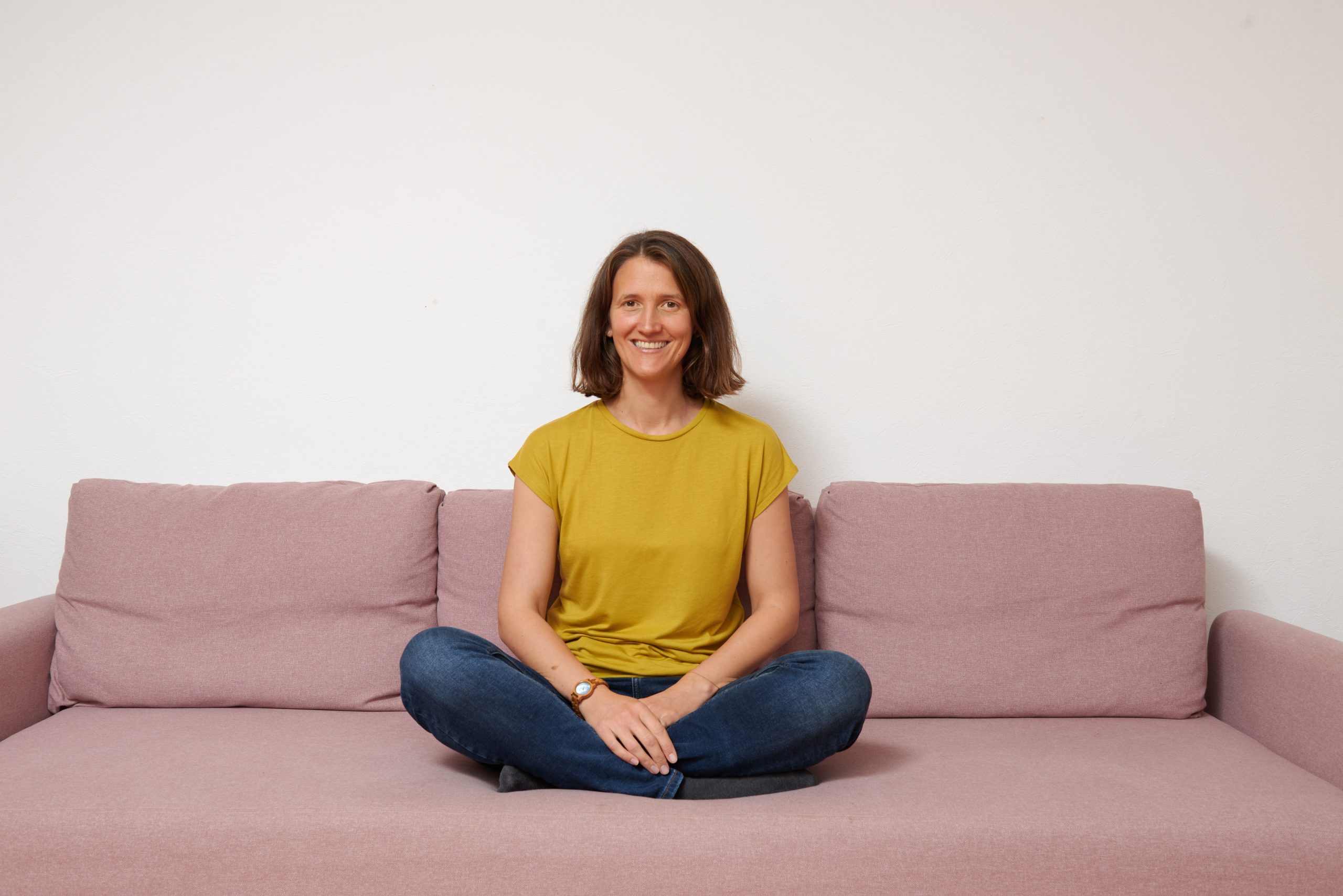 Portrait Jennifer Herrmann, Jennifer sitzt im Schneidersitz auf einem Sofa und blickt den Betrachter offen und einladend an. 